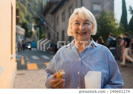 Happy senior woman tourist holding paper bag with pastry outdoors Happy senior woman tourist holding paper bag with pastry outdoors 140681274