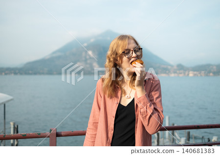 Beautiful young caucasian woman wearing eyeglasses looking at camera and enjoying a snack or pastry outside on a sunny summer day with beautiful backlighting. Beautiful young caucasian woman wearing eyeglasses looking at camera and enjoying a snack or pastry outside on a sunny summer day with beautiful backlighting. 140681383