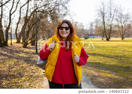 Happy woman in yellow vest and sunglasses walking along park path with backpack, enjoying sunny day and outdoor lifestyle. Happy woman in yellow vest and sunglasses walking along park path with backpack, enjoying sunny day and outdoor lifestyle. 140681522