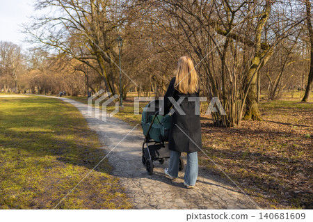 30s mother woman walking along park path while pushing baby stroller, enjoying sunny day and outdoor parenting lifestyle. 140681609