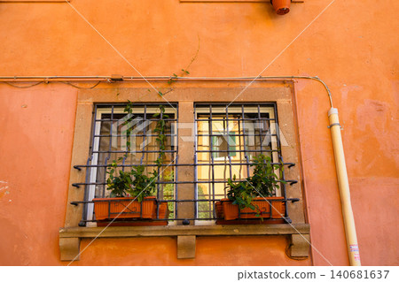 Classic rectangular window with decorative iron bars and flower pots on a bright orange stucco wall. Classic rectangular window with decorative iron bars and flower pots on a bright orange stucco wall. 140681637