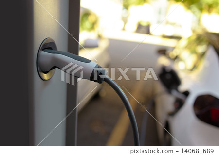 Close-up of electric car charging plug inserted into power station connector with vehicles in background. Close-up of electric car charging plug inserted into power station connector with vehicles in background. 140681689