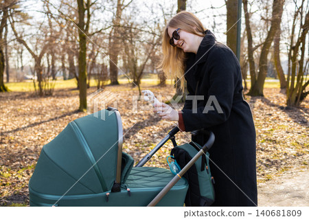 Mother holding baby toy while standing next to stroller in park, outdoor parenting and childcare lifestyle concept. 140681809