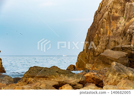 Beyond the desolate rocky shore, a calm sea and a single ship are visible. 140682005