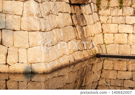 The stone walls of the castle illuminated by the setting sun and their inverted reflection on the water's surface. 140682007