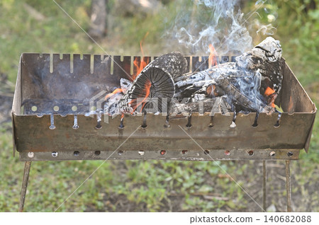 Burning firewood with smoke inside metal grill outdoors during summer picnic. Firewood glows with orange flames in charcoal brazier. Hot firewood prepares heat for barbecue in garden. Burning firewood with smoke inside metal grill outdoors during summer picnic. Firewood glows with orange flames in charcoal brazier. Hot firewood prepares heat for barbecue in garden. 140682088