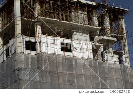 Construction building scaffolding high rise concrete renovation facade safety, unfinished structure wrapped with netting, sunlit exterior with window opening, urban development and progress 140682738
