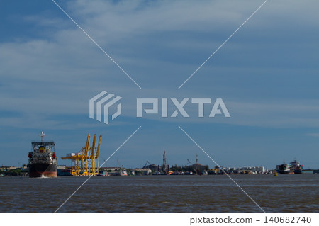 Port harbor cargo ship quay crane river shoreline sky view across brown water, industrial waterfront with loading equipment and distant vessel under calm cloud layer 140682740