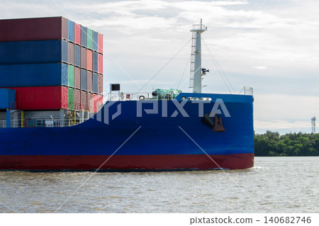 Container ship cargo shipping container maritime transport logistics freight vessel cruising along river under cloudy sky, blue hull beside stacked containers, global trade concept 140682746