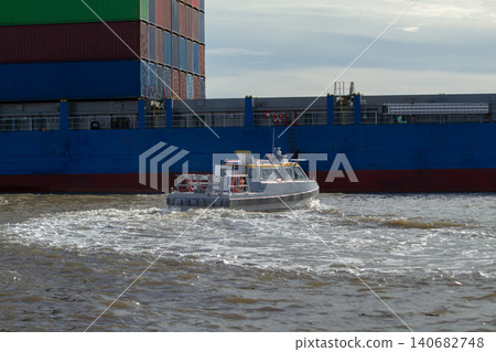 Pilot boat container ship cargo shipping harbor river maritime logistics, small service vessel cutting wake beside stacked freight container, industrial port transport scene under cloudy sky Pilot boat container ship cargo shipping harbor river maritime logistics, small service vessel cutting wake beside stacked freight container, industrial port transport scene under cloudy sky 140682748