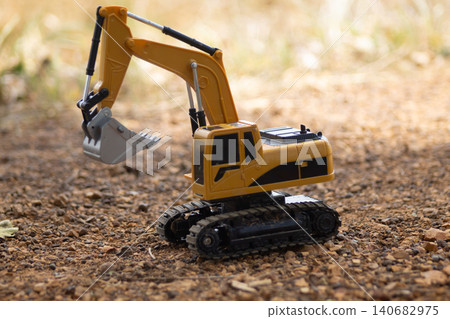 Toy excavator construction vehicle yellow track bucket gravel outdoor miniature equipment on dirt and pebble surface, closeup with sunlight bokeh, playful childhood building scene Toy excavator construction vehicle yellow track bucket gravel outdoor miniature equipment on dirt and pebble surface, closeup with sunlight bokeh, playful childhood building scene 140682975