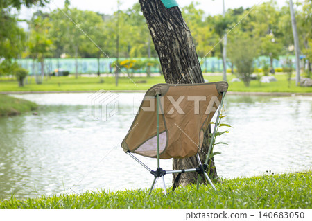 Folding chair by tree beside lake in green park, calm outdoor leisure scene with grass, water, and blurred trees Folding chair by tree beside lake in green park, calm outdoor leisure scene with grass, water, and blurred trees 140683050