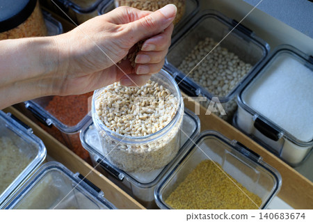Pouring pearl barley from woman hand into plastic container, kitchen pantry drawer storage with dry groats close up Pouring pearl barley from woman hand into plastic container, kitchen pantry drawer storage with dry groats close up 140683674