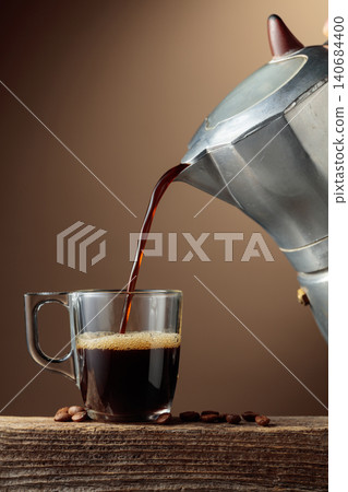 Black coffee is poured from a classic Italian aluminium moka pot into a glass coffee cup. 140684400
