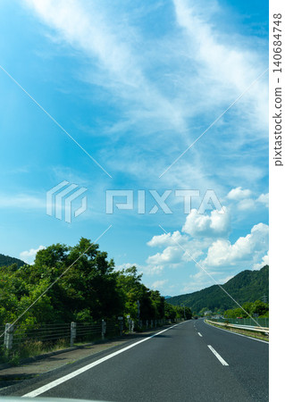 A view of a highway stretching across a blue sky and green mountains. A view of a highway stretching across a blue sky and green mountains. 140684748