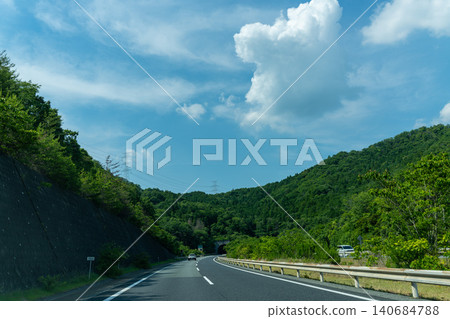 A view of a highway stretching across a blue sky and green mountains. A view of a highway stretching across a blue sky and green mountains. 140684788
