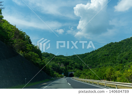 A view of a highway stretching across a blue sky and green mountains. A view of a highway stretching across a blue sky and green mountains. 140684789