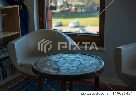 Worn white armchairs by window with metal table and EU flag indoors Worn white armchairs by window with metal table and EU flag indoors 140685434