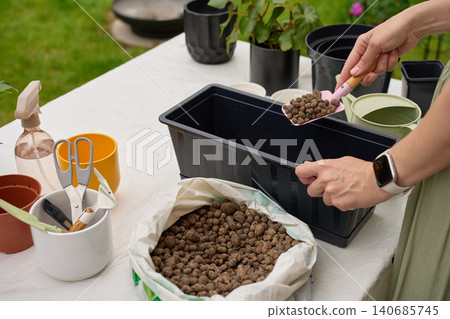 Woman adds clay pebbles to black planter box with hand trowel at an outdoor gardening table Woman adds clay pebbles to black planter box with hand trowel at an outdoor gardening table 140685745