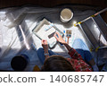Overhead view of person holding paint roller above tray with bucket, brushes and ladder on protective sheet 140685747