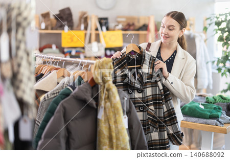 Positive young woman choosing winter coat in clothing store Positive young woman choosing winter coat in clothing store 140688092