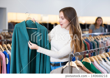 Positive young woman is thinking about buying a turquoise cardigan while walking through a clothing store Positive young woman is thinking about buying a turquoise cardigan while walking through a clothing store 140688218