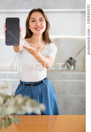 In kitchen, smiling girl in casual clothes shows blank smartphone screen close-up In kitchen, smiling girl in casual clothes shows blank smartphone screen close-up 140688432