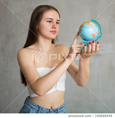 Young girl with unfastened hair stands with globe in her hands. Points finger at desired destination Young girl with unfastened hair stands with globe in her hands. Points finger at desired destination 140688444