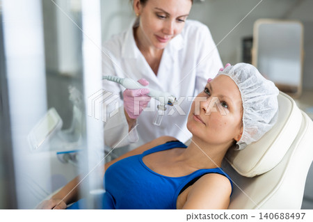 Young female cosmetologist doing moisturizing procedure to adult woman Young female cosmetologist doing moisturizing procedure to adult woman 140688497