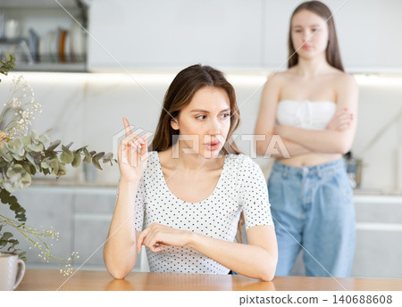 Angry mother sitting at the kitchen table, daughter standing behind Angry mother sitting at the kitchen table, daughter standing behind 140688608
