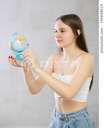 Young girl with unfastened hair stands with globe in her hands. Points finger at desired destination Young girl with unfastened hair stands with globe in her hands. Points finger at desired destination 140688619