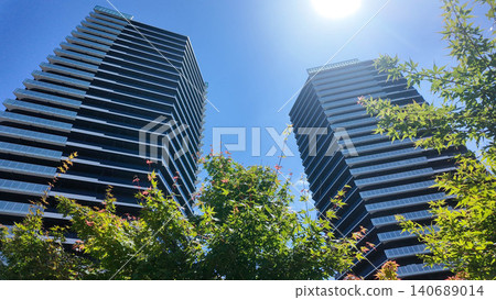 Low angle view of modern twin skyscraper buildings behind green trees and blue sky. Low angle view of modern twin skyscraper buildings behind green trees and blue sky. 140689014