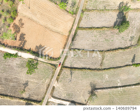 Harvested farmland with an orange tractor traveling along a rural dirt road. Agricultural field patterns, seasonal farm activity and countryside land management practices. Sustainable farming. 140689794