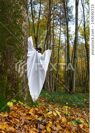 Playful ghost halloween costume White fabric drapes over the figure, while glasses and cap add personality charm. Colorful fall foliage tree trunks fallen leaves and natural woodland surroundings Playful ghost halloween costume White fabric drapes over the figure, while glasses and cap add personality charm. Colorful fall foliage tree trunks fallen leaves and natural woodland surroundings 140690318