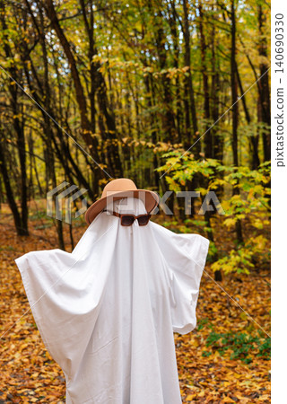 Playful ghost halloween costume White fabric drapes over the figure, while glasses and hat add personality charm. Colorful fall foliage tree trunks fallen leaves and natural woodland surroundings Playful ghost halloween costume White fabric drapes over the figure, while glasses and hat add personality charm. Colorful fall foliage tree trunks fallen leaves and natural woodland surroundings 140690330
