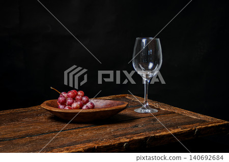 Fresh red grapes in wooden bowl and clear wine glass on rustic wooden table, representing flavor, gourmet appetizers, and elegant dark background simplicity Fresh red grapes in wooden bowl and clear wine glass on rustic wooden table, representing flavor, gourmet appetizers, and elegant dark background simplicity 140692684