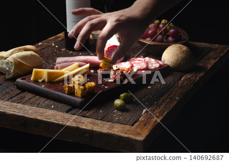 Hand picking flavorful appetizers from a rustic charcuterie board with sliced salami, cheeses, cured meats, bread, olives, and red wine bottle Hand picking flavorful appetizers from a rustic charcuterie board with sliced salami, cheeses, cured meats, bread, olives, and red wine bottle 140692687
