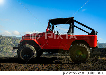 A red jeep stands on volcanic ground with hills in view A red jeep stands on volcanic ground with hills in view 140692841