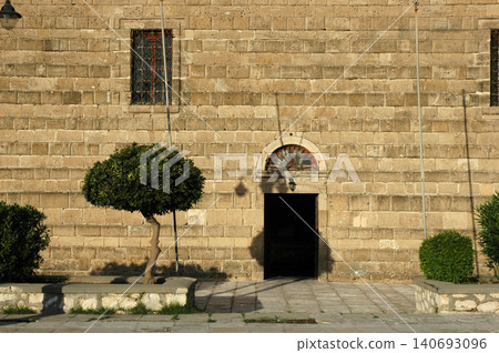 Historic stone facade of Saint Nicholas of the Molos Church in Zakynthos Town with a symmetrical view of the entrance and surrounding green shrubs 140693096