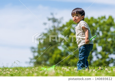 A boy runs across a grassy field under a blue sky, carrying a butterfly net. 140693612