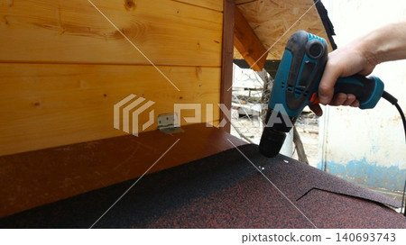 The process of outdoor construction or repair work involving the installation of roofing. A man's hand holds a corded or cordless screwdriver and screws a screw into a roof deck covered with dark roof 140693743
