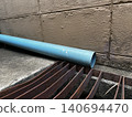 Blue PVC pipe resting on rusty metal grates against weathered concrete wall in industrial outdoor setting with rough textured surfaces 140694470