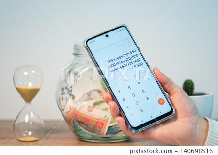 Counting saved money for a purchase. Phone with calculator in female hands with jar with saved up money on the background Counting saved money for a purchase. Phone with calculator in female hands with jar with saved up money on the background 140698516