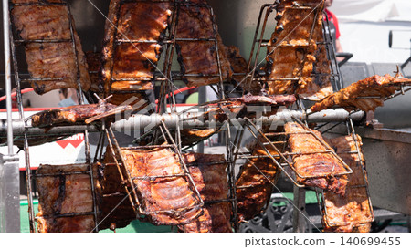 Rows of meat racks are cooking over open heat on a metal barbecue setup with browned surfaces, charred edges, and an outdoor food preparation background 140699455