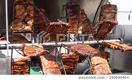 Several slabs of meat are cooking on metal racks over an open grill setup, with dark charred surfaces, warm browning, and industrial cooking equipment visible in the background Several slabs of meat are cooking on metal racks over an open grill setup, with dark charred surfaces, warm browning, and industrial cooking equipment visible in the background 140699464