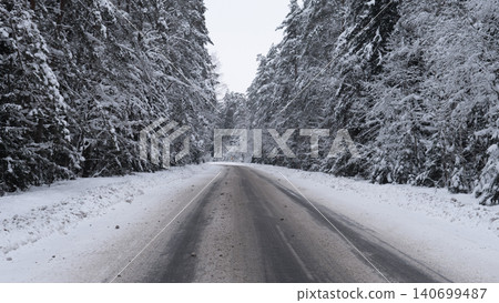 Snow covered two-lane road stretching into distance between tall evergreen trees heavily laden with fresh snow, roadside snowbanks and scattered slush on 140699487