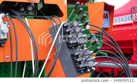 Detailed close-up of an orange and green agricultural machine with hydraulic control levers, black hoses, metal fittings, and mechanical components in an outdoor farm setting Detailed close-up of an orange and green agricultural machine with hydraulic control levers, black hoses, metal fittings, and mechanical components in an outdoor farm setting 140699512