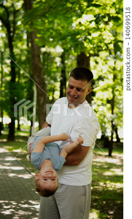 Man holding a laughing little boy upside down while walking along a tree-lined park path; dappled sunlight filters through leafy canopy creating soft 140699518