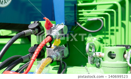 Close-up view of bright green industrial machinery showing metal connectors, thick hoses, colored cables, and mechanical fittings with strong daylight and blurred equipment in 140699523