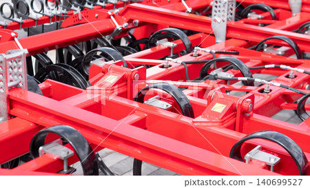 Close-up view of bright red agricultural machinery parts with black wheels, metal brackets, cables, and mechanical assemblies arranged in rows under even industrial lighting Close-up view of bright red agricultural machinery parts with black wheels, metal brackets, cables, and mechanical assemblies arranged in rows under even industrial lighting 140699527